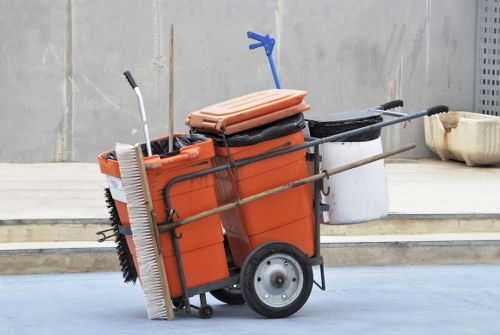 A mobile rubbish collection cart positioned outdoors on a paved surface, used for waste removal services in Battersea. The cart features an orange plastic bin with a black lid, mounted on a metal frame with two large rubber wheels at the back and legs at the front for stability. Attached to the frame are various cleaning and sweeping tools, including a large push broom with white bristles leaning against the side, a hand broom with black bristles, and a long-handled tool possibly for lifting trash. Resting on top of the bin is a folded orange container, and a blue plastic hook is attached to the top of the bin frame. The cart is set against a plain concrete wall with a narrow step leading up to a courtyard or alleyway, suggesting a typical urban setting for waste collection. This image visually represents waste management activities associated with commercial rubbish clearance services, like those provided by Commercial Waste Removal Battersea, within the Wandsworth area.