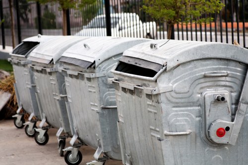 Four large, silver metal rubbish bins with curved lids and sturdy wheels are lined up on a paved surface, likely in a commercial or residential area near Battersea. The bins are positioned side by side, with the first bin in the foreground and the others receding into the background, slightly angled to the left. The lids are closed, with the front of the lids slightly raised, revealing openings for waste disposal. The surface of the bins shows signs of weathering with minor scratches and spots. Behind the bins, there is a black metal fence and some greenery, including trees and bushes, indicating an outdoor environment in a town setting. The overall scene suggests a typical rubbish collection point managed by a waste removal service, such as Commercial Waste Removal Battersea, situated within a urban context in London postcodes around SW11 or SW8. The lighting appears natural, likely daytime, highlighting the metallic texture and functional design of the bins, which are essential for waste management and rubbish disposal in the local area.