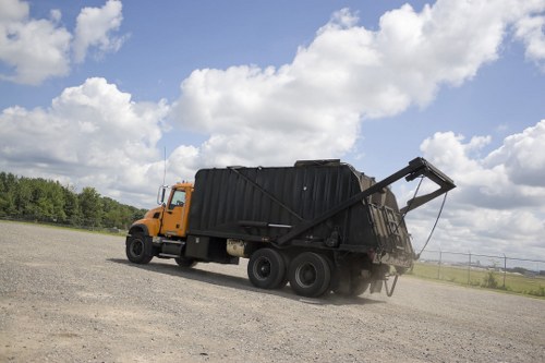 A large yellow and black waste collection truck is parked on an expansive, flat gravel surface under a partly cloudy sky. The truck's cab and front section are painted yellow, while the rear container for rubbish is black. The rear loading mechanism is extended outward at an angle, indicating that the container is being emptied or loaded. The vehicle is positioned in an open, outdoor environment with a rural landscape visible in the background, including distant trees and low-lying greenery. The scene suggests a commercial rubbish removal operation in a countryside or suburban setting, relevant to services such as those offered by Commercial Waste Removal Battersea for waste collection and disposal across London and surrounding areas.