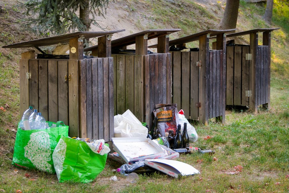 A collection of overflowing rubbish bins and scattered waste on a paved area in an urban setting, with cardboard boxes, plastic bags, and paper debris spilling onto the ground surrounding a large grey