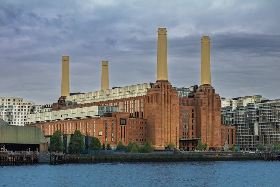 A view across the River Thames showing a large industrial-style power station with three tall chimneys made of red brick, situated on the opposite bank. In the foreground, part of a multi-storey struc