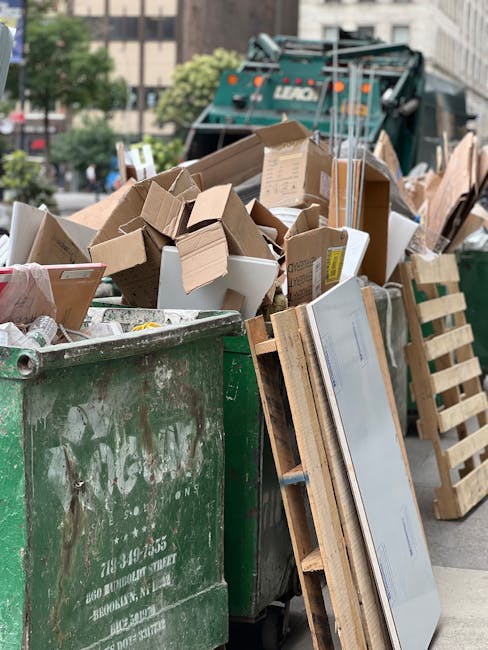 A row of large, wheeled rubbish bins in tandem along a pavement, with green, red, blue, and gray lids, each fitted with ridged, durable plastic surfaces. The bins are positioned closely together, some